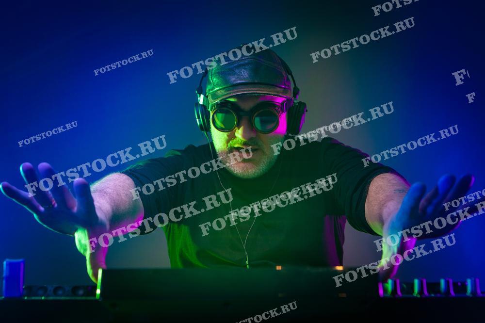 На изображении виден DJ за работой: он в наушниках, очках и с кепкой, руки раскинуты над микшерным пультом, а лицо и руки подсвечены в зелёном и синем цвете. Такая фотография может быть использована для рекламы музыкальных мероприятий, DJ-сервисов или в статьях о ночной жизни и клубной культуре.